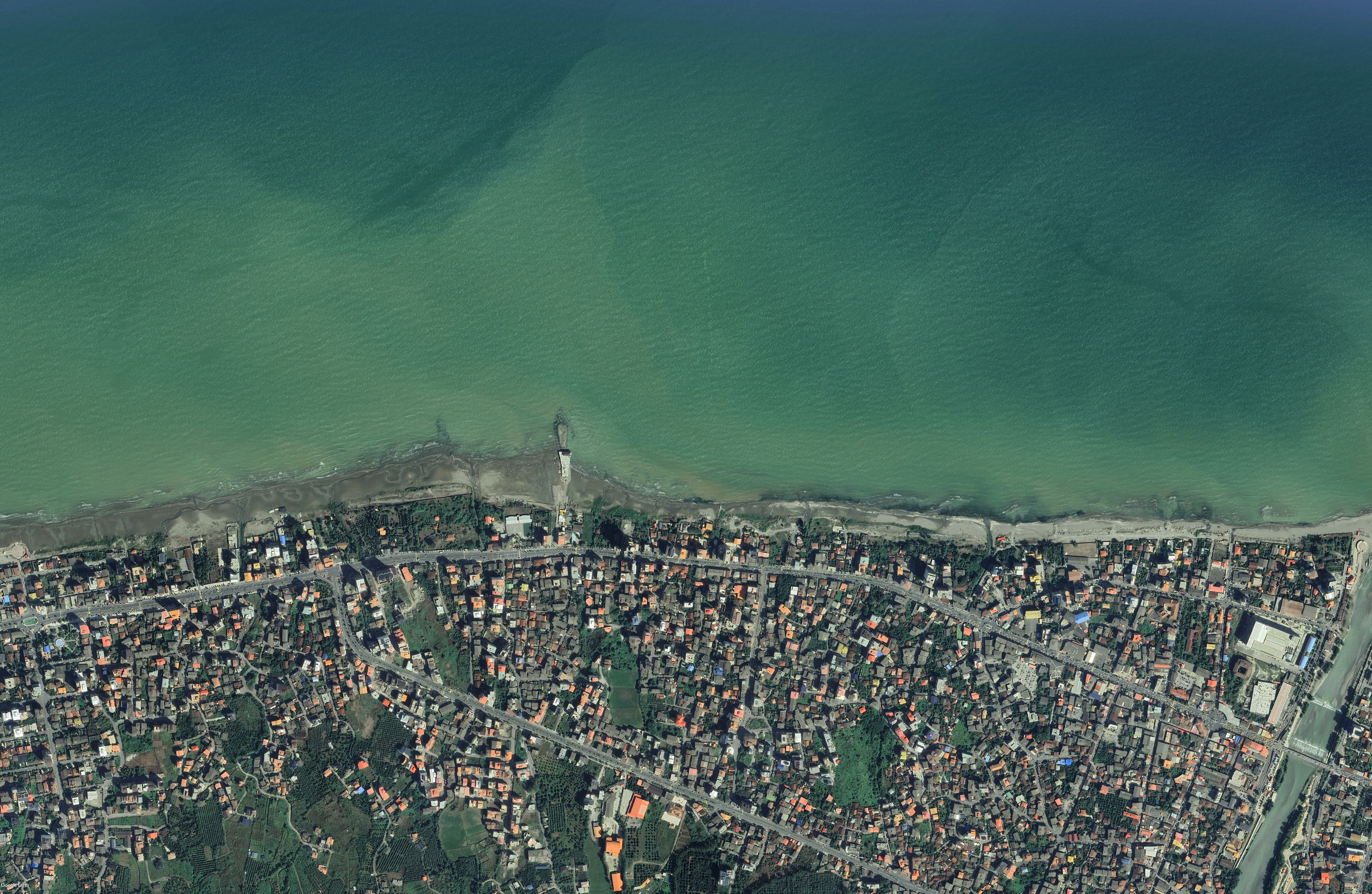 Wide satellite view of the coast, the tower alone on the shoreline with the town spread inland behind it